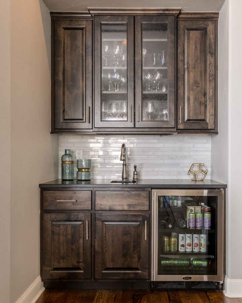 This is an example of a large transitional galley home bar in Denver with dark hardwood floors, marble splashback, a drop-in sink, raised-panel cabinets, dark wood cabinets, soapstone benchtops and white splashback.