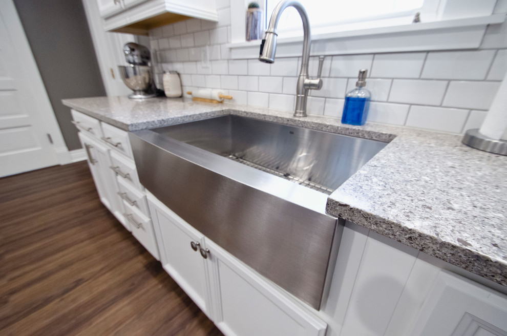 This is an example of a small country l-shaped separate kitchen in Chicago with a farmhouse sink, recessed-panel cabinets, white cabinets, quartz benchtops, white splashback, ceramic splashback, stainless steel appliances, vinyl floors, no island, brown floor and grey benchtop.