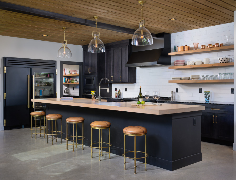 Foto på ett industriellt kök, med luckor med infälld panel, svarta skåp, träbänkskiva, vitt stänkskydd, stänkskydd i tunnelbanekakel, svarta vitvaror, betonggolv och en köksö