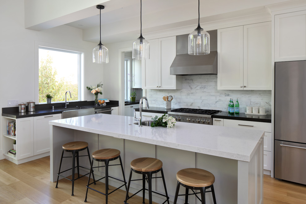 This is an example of a large country l-shaped eat-in kitchen in San Francisco with a farmhouse sink, shaker cabinets, white cabinets, white splashback, stainless steel appliances, medium hardwood floors, with island, brown floor, black benchtop, laminate benchtops and marble splashback.