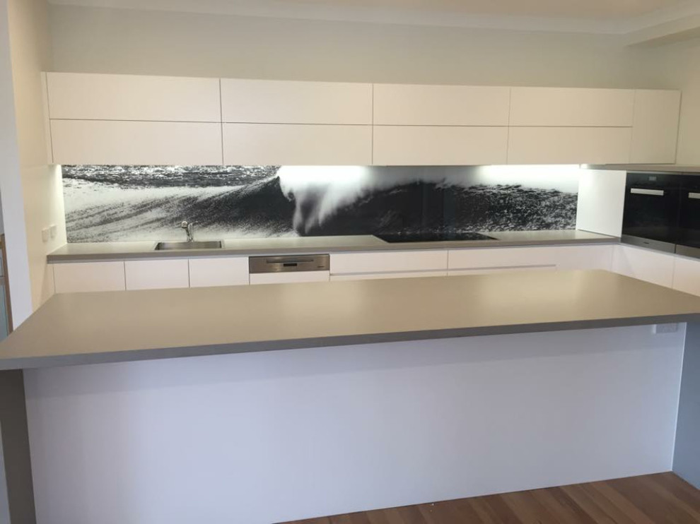 This is an example of a small contemporary single-wall eat-in kitchen in Sydney with an undermount sink, multi-coloured splashback, glass sheet splashback, stainless steel appliances, with island and brown floor.