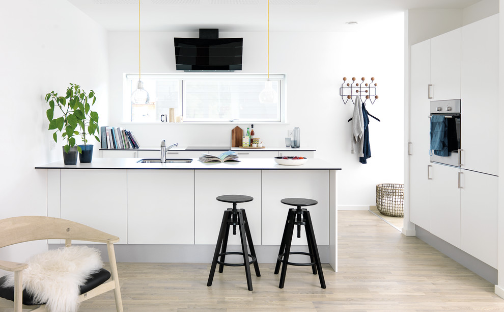 Mid-sized scandinavian galley open plan kitchen in Aarhus with an integrated sink, flat-panel cabinets, white cabinets, laminate benchtops, stainless steel appliances, light hardwood floors and a peninsula.
