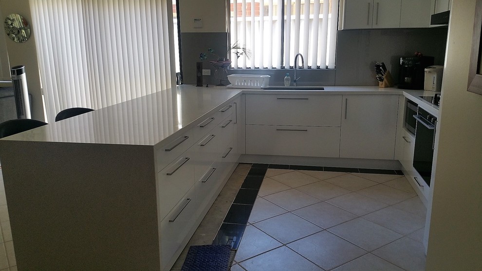 This is an example of a large contemporary u-shaped eat-in kitchen in Perth with a drop-in sink, flat-panel cabinets, white cabinets, quartz benchtops, grey splashback, glass sheet splashback, stainless steel appliances, ceramic floors, with island, white floor and white benchtop.
