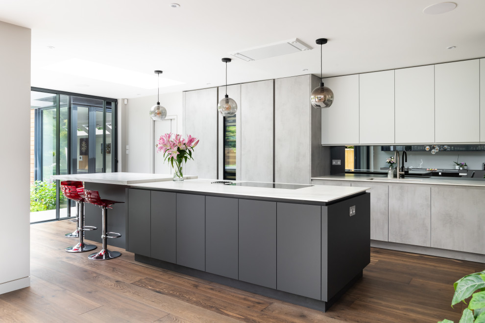 This is an example of an expansive modern l-shaped open plan kitchen in Surrey with a drop-in sink, grey cabinets, quartzite benchtops, mirror splashback, stainless steel appliances, medium hardwood floors, with island, brown floor and white benchtop.