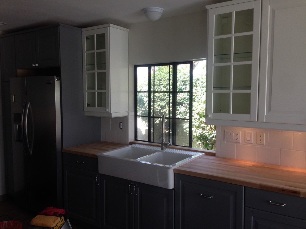 Example of a transitional u-shaped laminate floor and brown floor enclosed kitchen design with a farmhouse sink, glass-front cabinets, white cabinets, wood countertops, white backsplash and stainless steel appliances