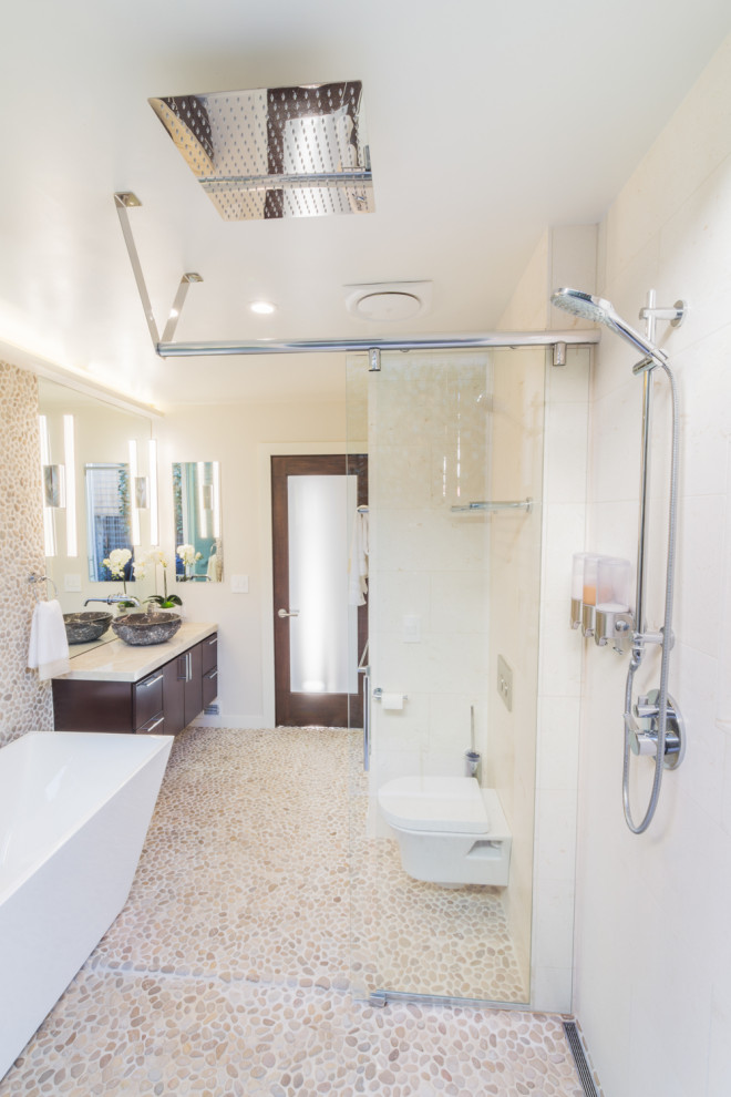 This is an example of a small contemporary master bathroom in Los Angeles with flat-panel cabinets, brown cabinets, a freestanding tub, a curbless shower, a wall-mount toilet, beige tile, pebble tile, beige walls, terrazzo floors, a vessel sink, marble benchtops, beige floor, a sliding shower screen and beige benchtops.