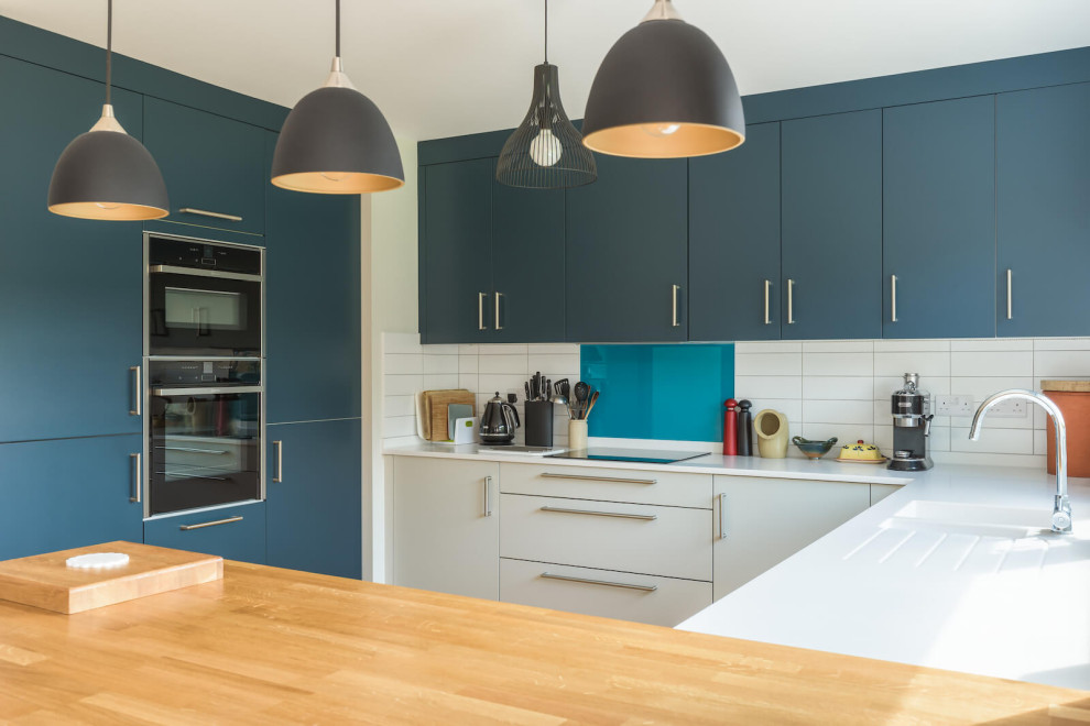 This is an example of a mid-sized traditional u-shaped open plan kitchen in Sussex with flat-panel cabinets, quartzite benchtops, white splashback, ceramic splashback, black appliances, with island and white benchtop.