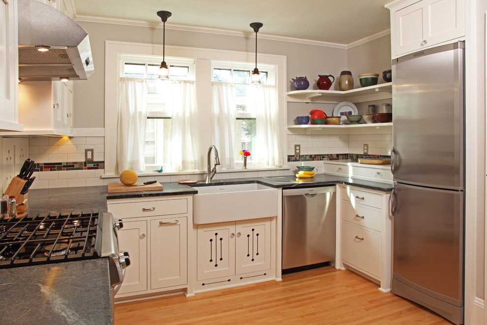 This is an example of a small arts and crafts l-shaped eat-in kitchen in Minneapolis with a farmhouse sink, recessed-panel cabinets, white cabinets, multi-coloured splashback, mosaic tile splashback, stainless steel appliances, light hardwood floors, no island, soapstone benchtops and beige floor.