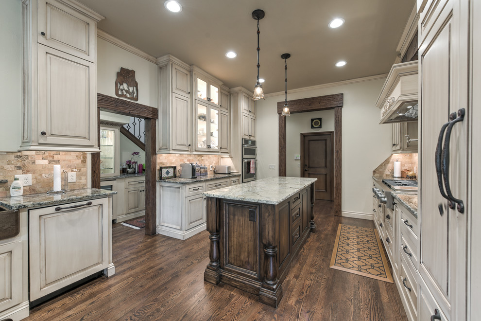 Photo of an expansive traditional galley separate kitchen in Oklahoma City with a farmhouse sink, raised-panel cabinets, light wood cabinets, granite benchtops, brown splashback, travertine splashback, stainless steel appliances, dark hardwood floors, with island and brown floor.
