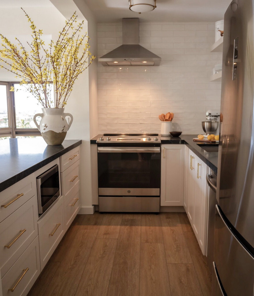 Small modern u-shaped eat-in kitchen in Hawaii with a drop-in sink, shaker cabinets, white cabinets, soapstone benchtops, white splashback, subway tile splashback, stainless steel appliances, vinyl floors, with island, brown floor and black benchtop.