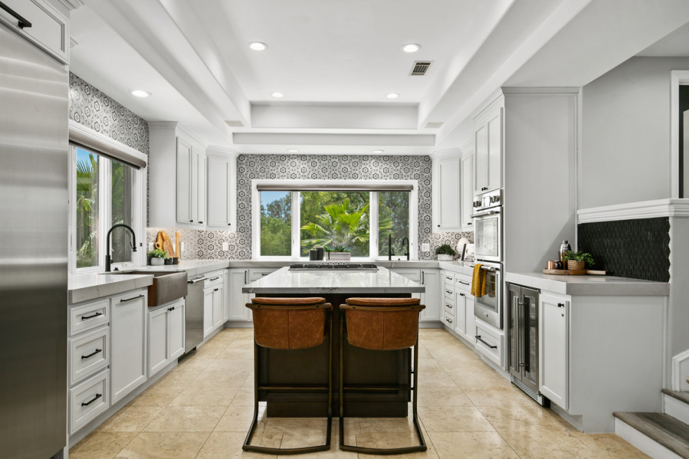 Inspiration for a large transitional u-shaped limestone floor, beige floor and vaulted ceiling open concept kitchen remodel in Orange County with a farmhouse sink, recessed-panel cabinets, white cabinets, marble countertops, multicolored backsplash, porcelain backsplash, stainless steel appliances, an island and gray countertops