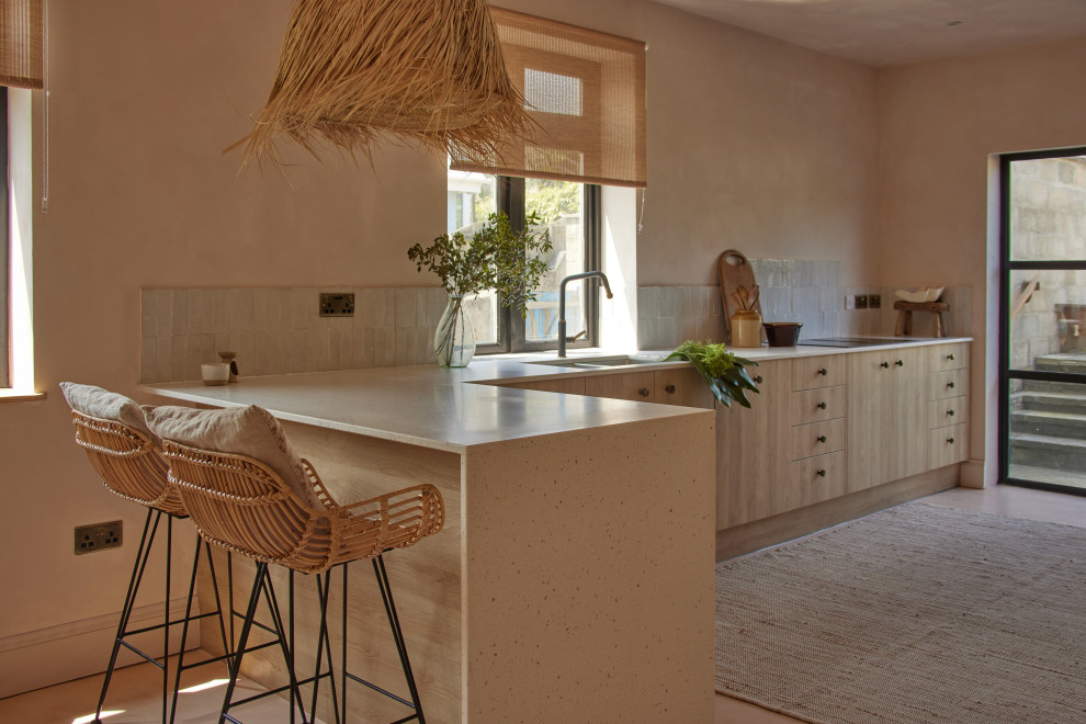 Inspiration for a large mediterranean l-shaped eat-in kitchen in Cornwall with an undermount sink, flat-panel cabinets, light wood cabinets, solid surface benchtops, beige splashback, ceramic splashback, black appliances, concrete floors, a peninsula, pink floor and beige benchtop.