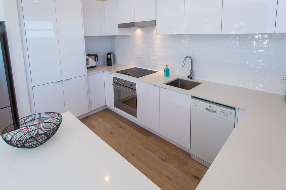 Photo of a small beach style u-shaped eat-in kitchen in Sydney with a drop-in sink, flat-panel cabinets, white cabinets, quartz benchtops, white splashback, subway tile splashback, stainless steel appliances, light hardwood floors and no island.