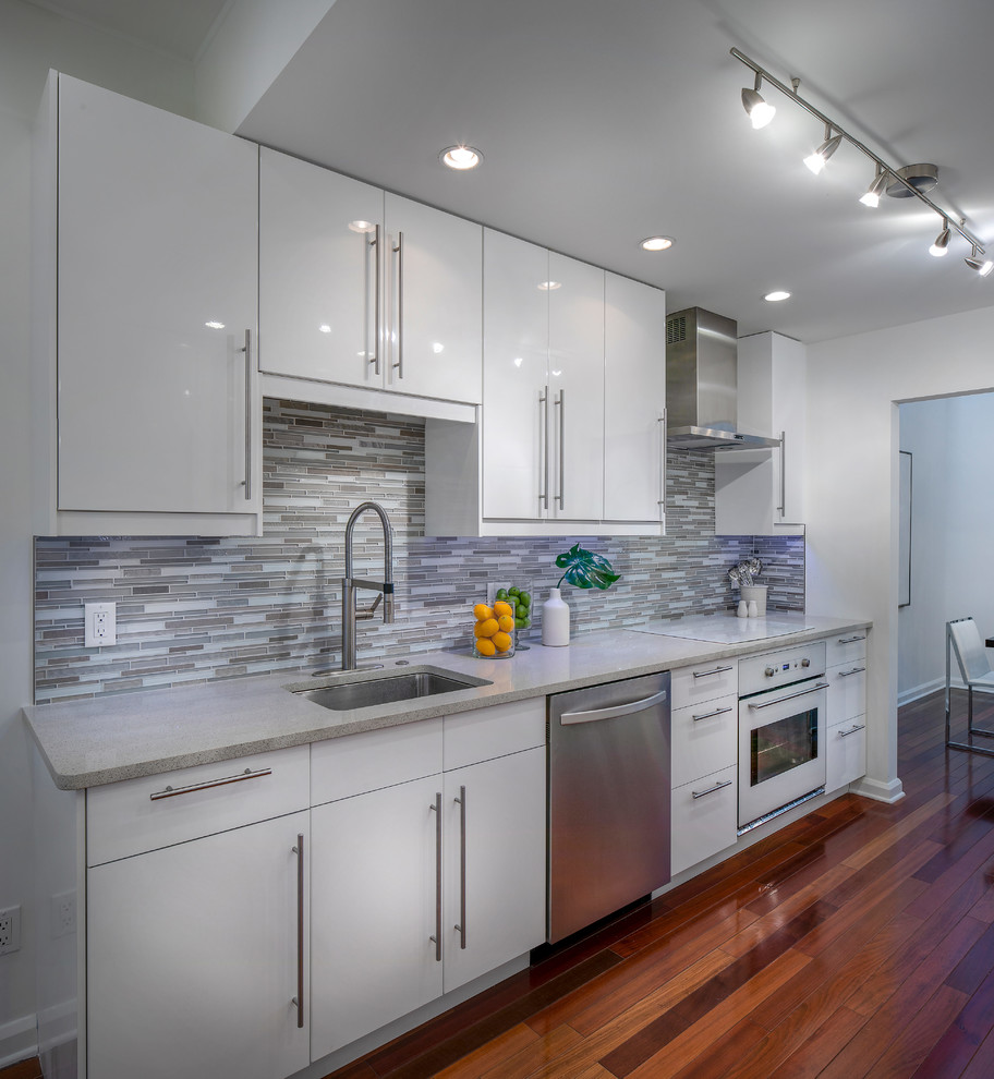 Inspiration for a small modern galley kitchen in Tampa with an undermount sink, flat-panel cabinets, white cabinets, quartz benchtops, grey splashback, glass tile splashback, stainless steel appliances, dark hardwood floors, brown floor and grey benchtop.