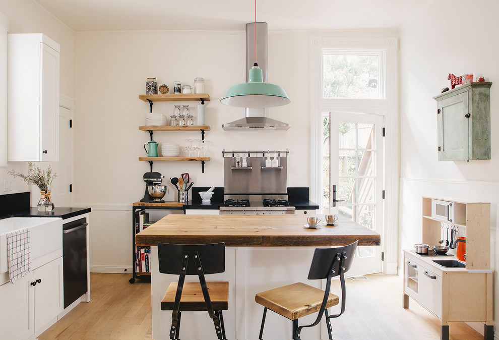 Photo of a scandinavian kitchen in San Francisco with a farmhouse sink, recessed-panel cabinets, white cabinets, wood benchtops, black splashback, stone slab splashback, stainless steel appliances, light hardwood floors and with island.