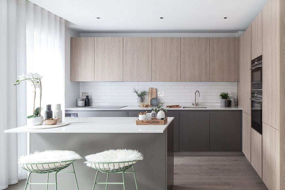Contemporary l-shaped kitchen in London with a submerged sink, flat-panel cabinets, light wood cabinets, white splashback, metro tiled splashback, black appliances, a breakfast bar, grey floors and white worktops.