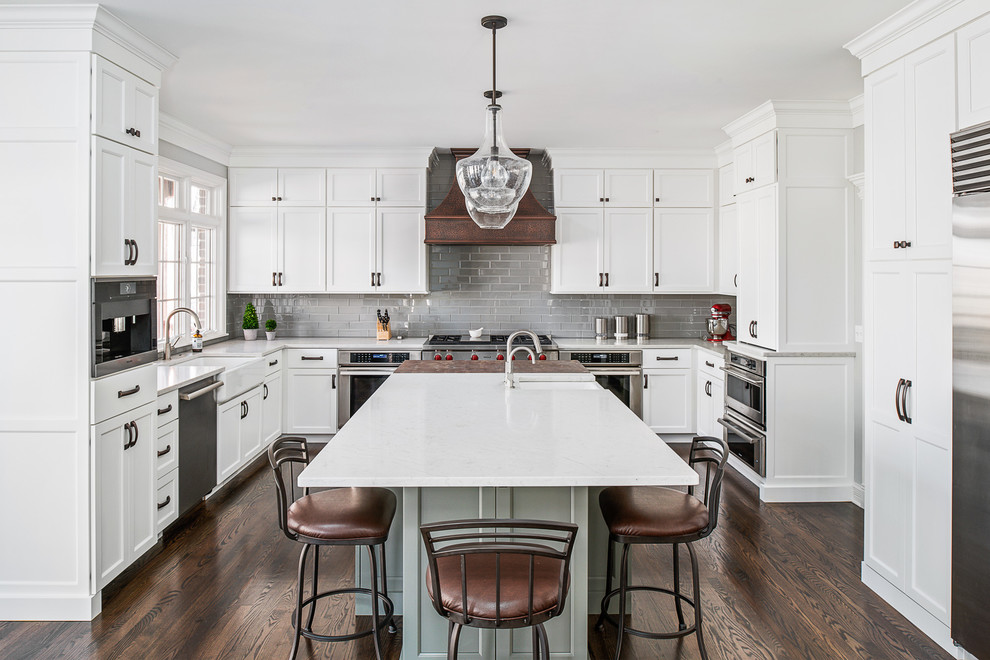 Photo of a large traditional u-shaped kitchen in Chicago with a farmhouse sink, white cabinets, quartz benchtops, grey splashback, glass tile splashback, stainless steel appliances, dark hardwood floors, with island, brown floor, white benchtop and shaker cabinets.