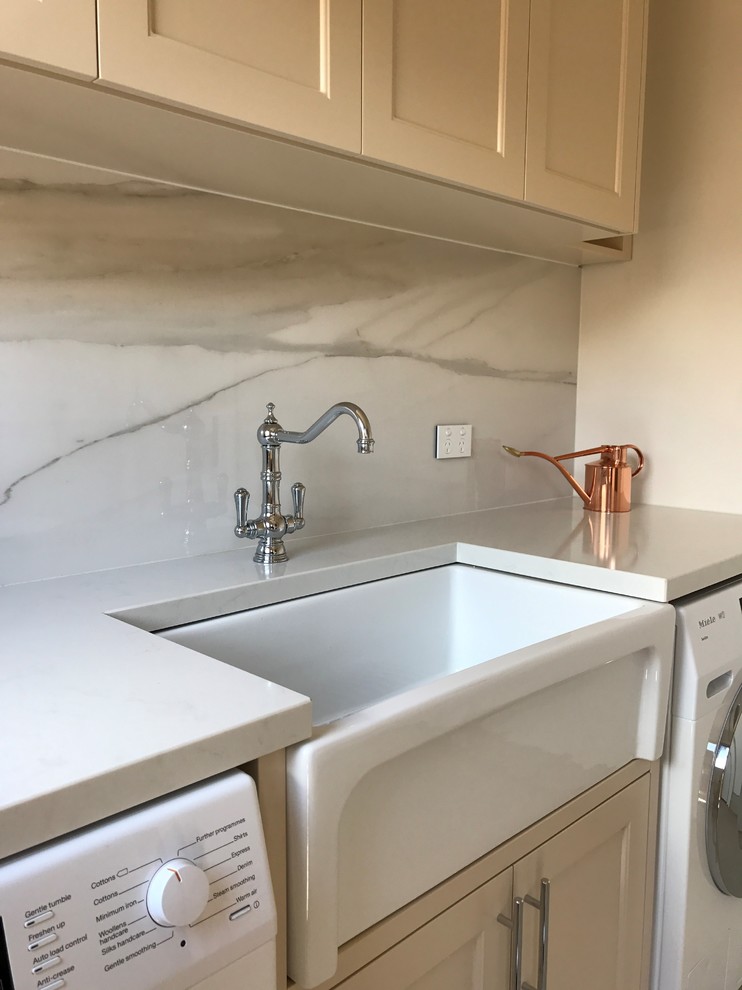 This is an example of a mid-sized modern galley dedicated laundry room in Sydney with shaker cabinets, beige cabinets, beige walls, porcelain floors, a farmhouse sink, quartz benchtops and a side-by-side washer and dryer.