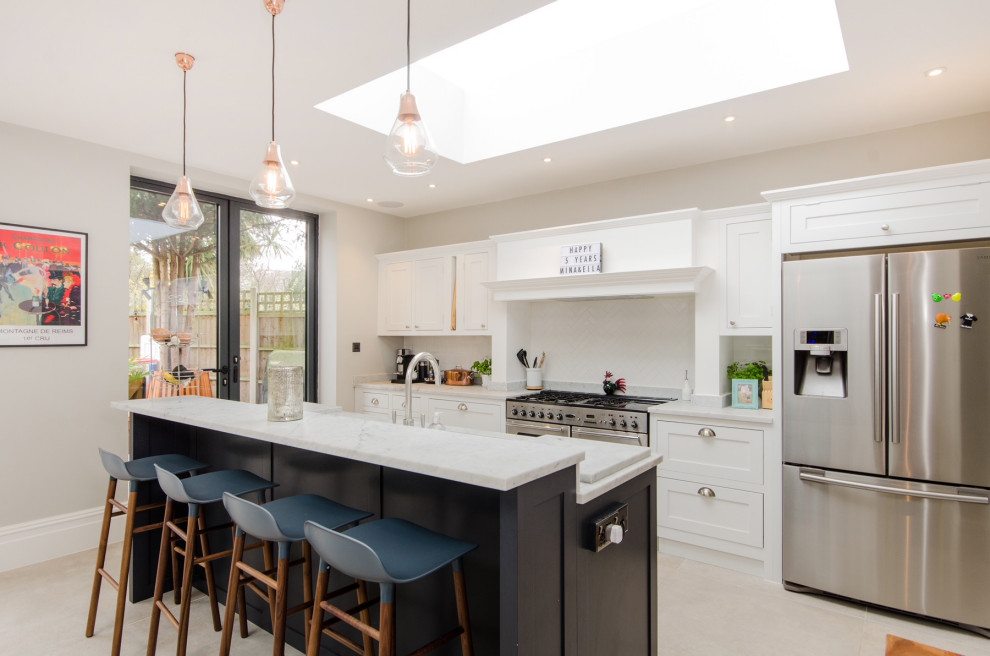 Photo of a transitional galley eat-in kitchen in London with shaker cabinets, white cabinets, white splashback, stainless steel appliances, with island, white floor and white benchtop.