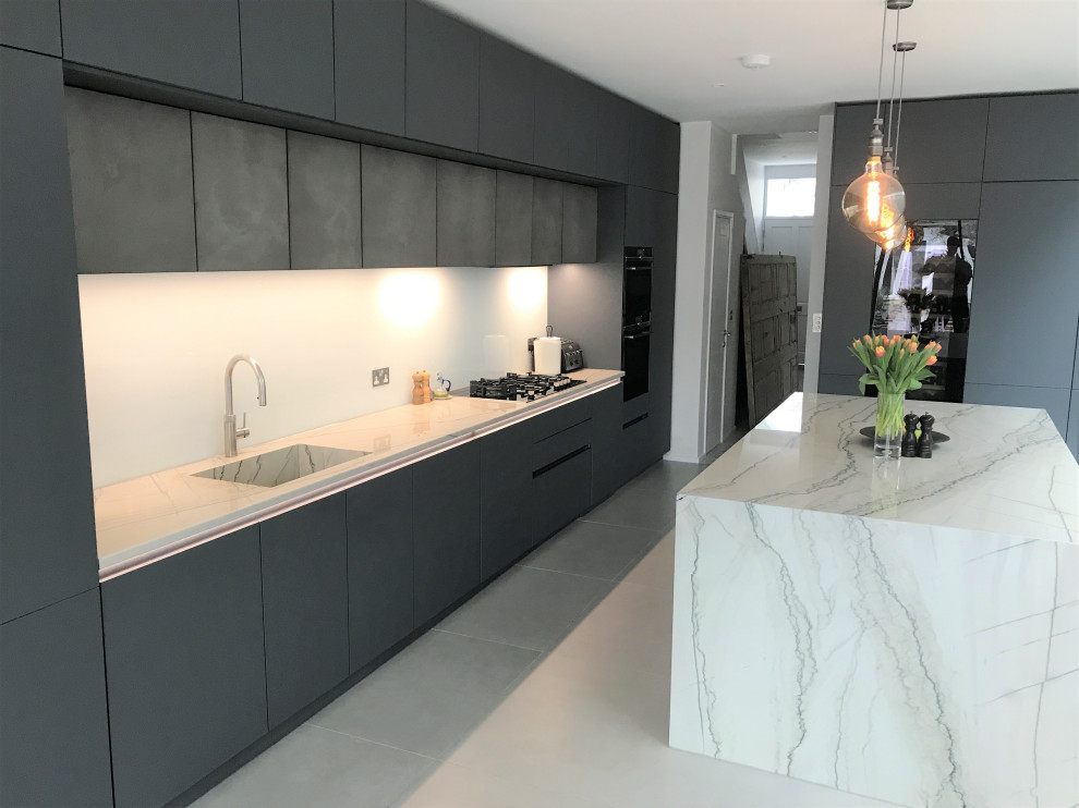 Photo of a large contemporary l-shaped eat-in kitchen in London with an undermount sink, flat-panel cabinets, grey cabinets, quartzite benchtops, white splashback, glass sheet splashback, black appliances, ceramic floors, with island, grey floor and grey benchtop.