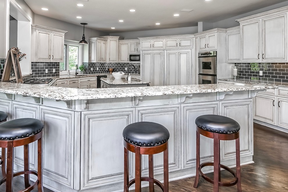 Large beach style u-shaped eat-in kitchen in Omaha with an undermount sink, recessed-panel cabinets, white cabinets, granite benchtops, grey splashback, glass tile splashback, stainless steel appliances, dark hardwood floors and with island.
