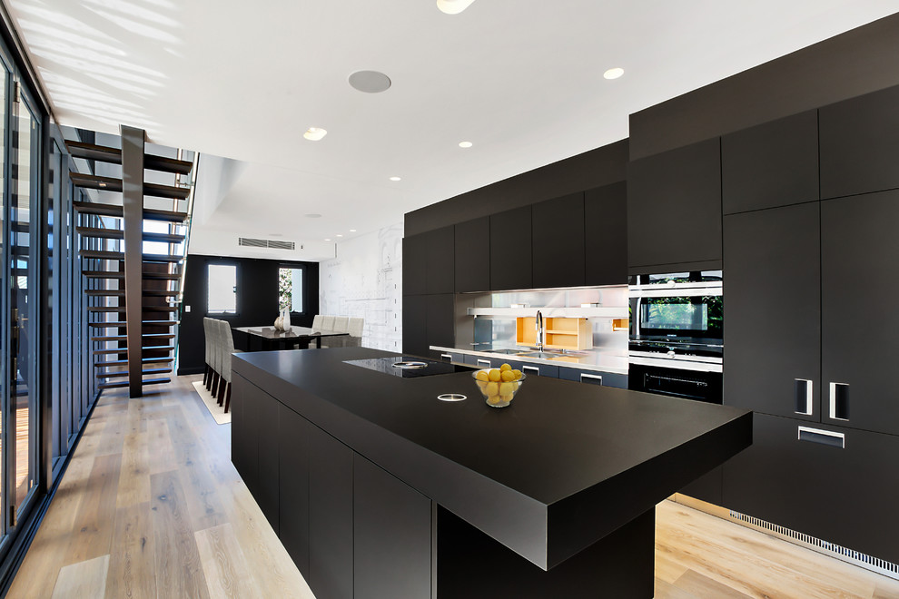 Photo of a contemporary galley eat-in kitchen in Sydney with an integrated sink, flat-panel cabinets, black cabinets, metallic splashback, black appliances, light hardwood floors, with island and black benchtop.