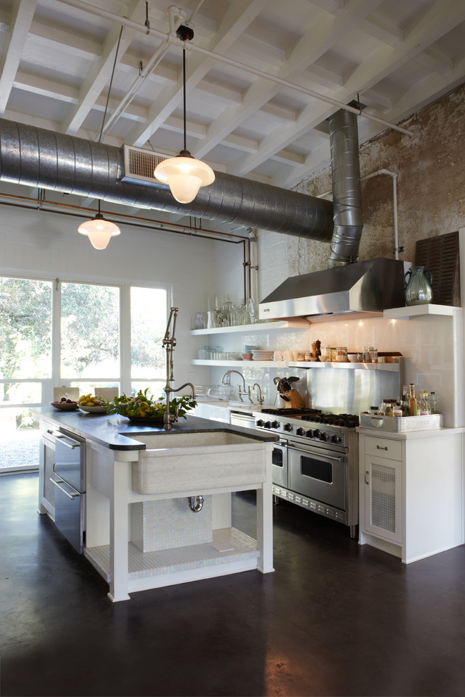 This is an example of a large industrial single-wall open plan kitchen in Orange County with a farmhouse sink, glass-front cabinets, white cabinets, solid surface benchtops, metallic splashback, metal splashback, stainless steel appliances, concrete floors and with island.