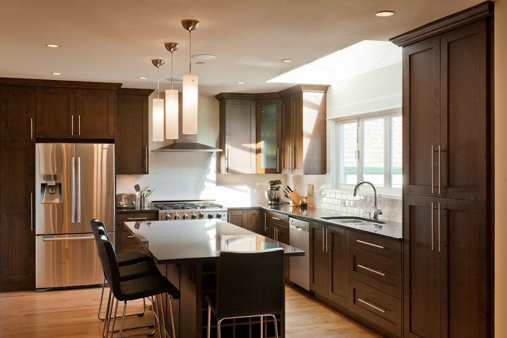Photo of a mid-sized contemporary l-shaped eat-in kitchen in Vancouver with subway tile splashback, stainless steel appliances, a double-bowl sink, shaker cabinets, dark wood cabinets, light hardwood floors, with island, brown floor, white splashback, granite benchtops and black benchtop.