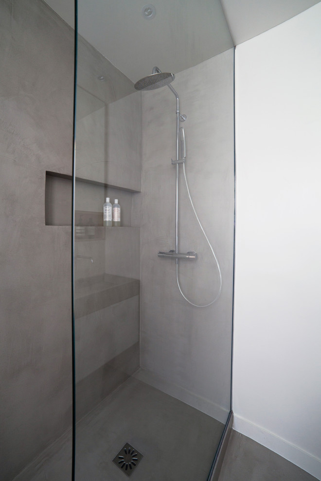 Inredning av ett mellanstort grå grått badrum med dusch, med luckor med profilerade fronter, vita skåp, en kantlös dusch, en vägghängd toalettstol, grå kakel, vita väggar, betonggolv, ett avlångt handfat, bänkskiva i betong, grått golv och dusch med gångjärnsdörr