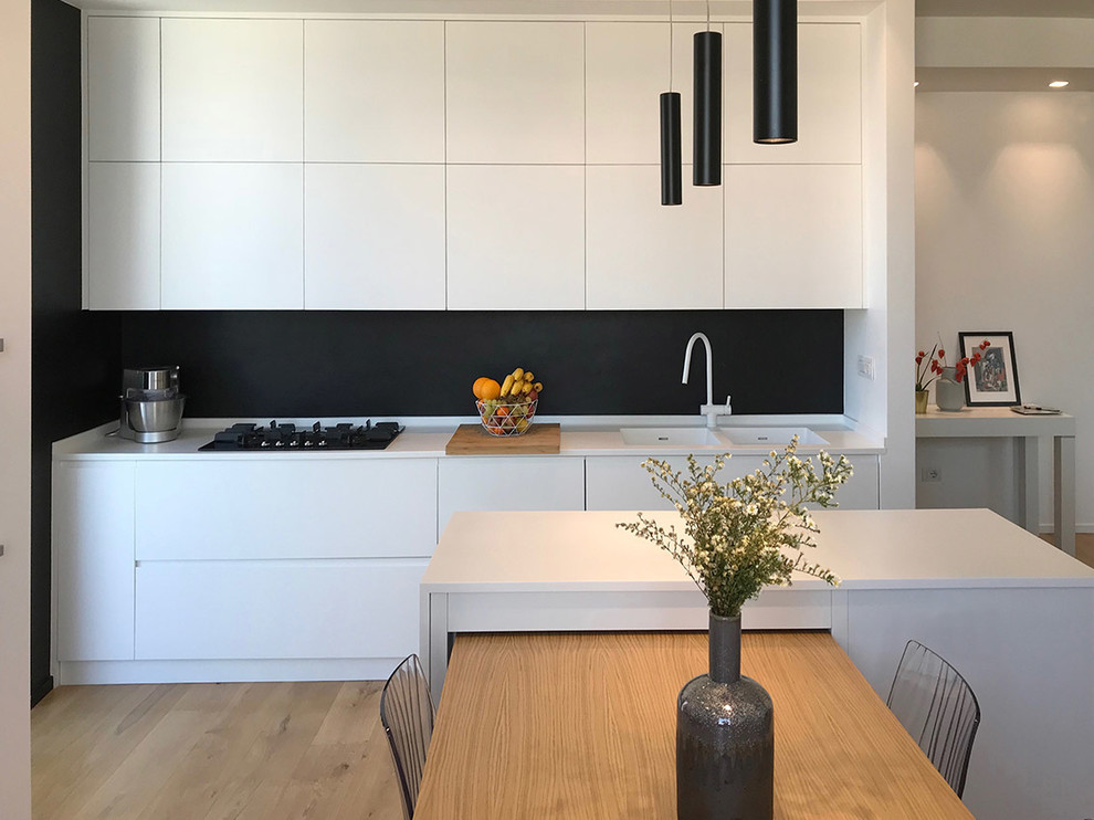 This is an example of a mid-sized contemporary single-wall eat-in kitchen in Rome with flat-panel cabinets, white cabinets, black splashback, a double-bowl sink, light hardwood floors, solid surface benchtops, stainless steel appliances, with island and white benchtop.