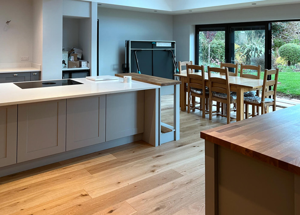 Photo of a large traditional grey and white u-shaped open plan kitchen in Sussex with a double-bowl sink, shaker cabinets, grey cabinets, quartz worktops, white splashback, black appliances, light hardwood flooring, an island, brown floors and white worktops.