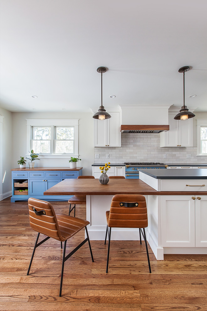 Inspiration for a medium sized classic l-shaped kitchen/diner in Detroit with a belfast sink, flat-panel cabinets, white cabinets, engineered stone countertops, white splashback, ceramic splashback, stainless steel appliances, medium hardwood flooring, an island, brown floors and black worktops.