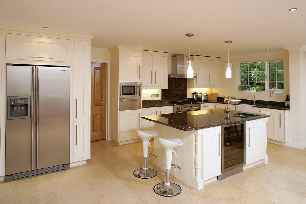 Photo of a large modern l-shaped eat-in kitchen in Hampshire with a farmhouse sink, shaker cabinets, white cabinets, granite benchtops, brown splashback, stone slab splashback, stainless steel appliances, limestone floors, with island, beige floor and brown benchtop.