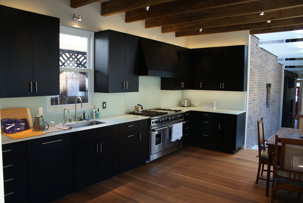 Inspiration for a mid-sized modern l-shaped eat-in kitchen in Denver with an undermount sink, flat-panel cabinets, black cabinets, glass benchtops, white splashback, glass sheet splashback, stainless steel appliances, dark hardwood floors and no island.