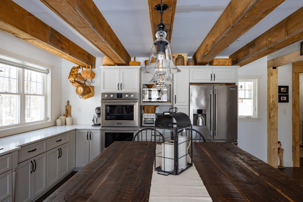 Photo of a large rural l-shaped kitchen/diner in Portland Maine with a belfast sink, shaker cabinets, grey cabinets, marble worktops, white splashback, tonge and groove splashback, stainless steel appliances, dark hardwood flooring, an island, brown floors, white worktops and exposed beams.