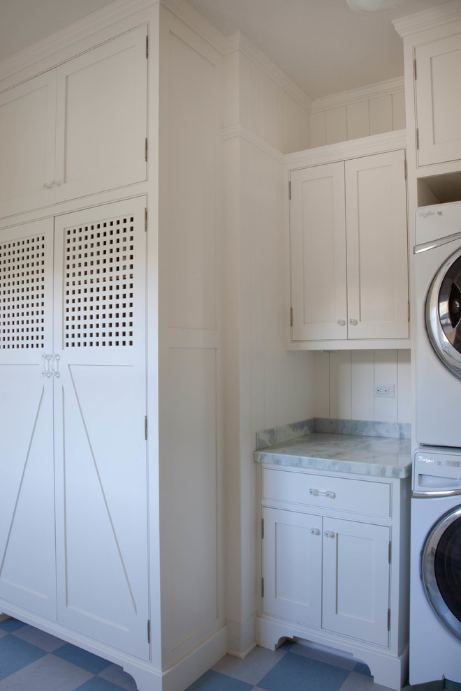 This is an example of a beach style galley dedicated laundry room in San Diego with a farmhouse sink, shaker cabinets, white cabinets, marble benchtops, white walls and a stacked washer and dryer.