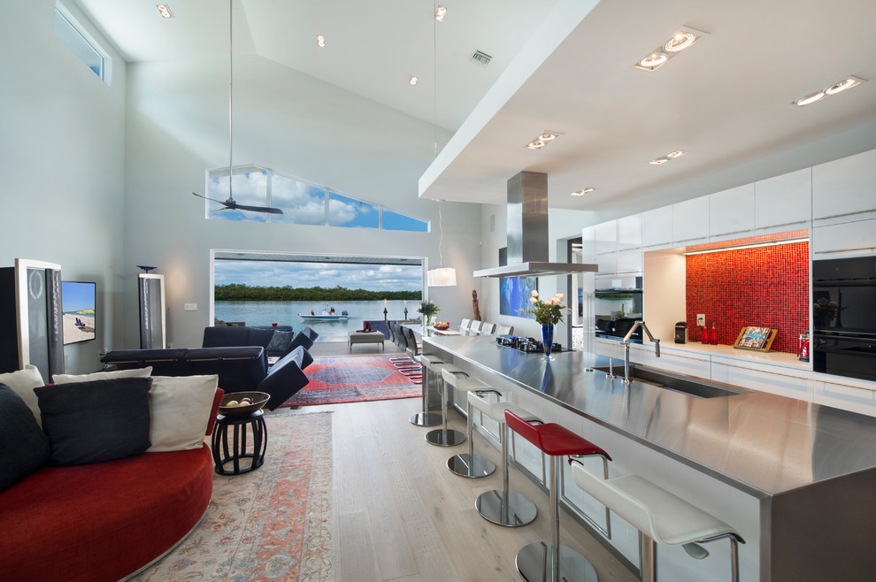 Design ideas for a large contemporary galley open plan kitchen in Other with light hardwood floors, flat-panel cabinets, white cabinets, red splashback, black appliances, with island, stainless steel benchtops, an integrated sink and glass tile splashback.