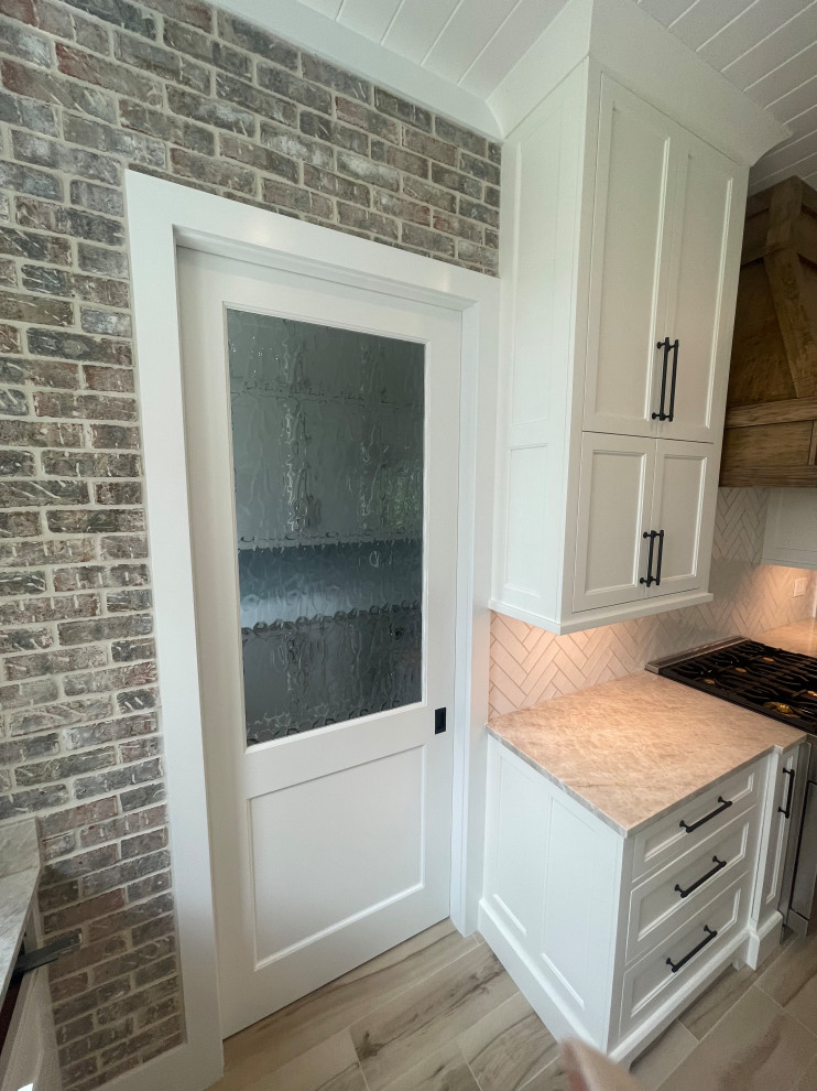 Photo of a mid-sized country u-shaped open plan kitchen in Orlando with a farmhouse sink, shaker cabinets, white cabinets, quartzite benchtops, white splashback, brick splashback, stainless steel appliances, ceramic floors, with island, beige floor, beige benchtop and timber.