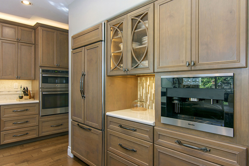 Large transitional l-shaped medium tone wood floor and brown floor open concept kitchen photo in San Diego with a farmhouse sink, recessed-panel cabinets, medium tone wood cabinets, beige backsplash, paneled appliances, an island, quartz countertops and travertine backsplash