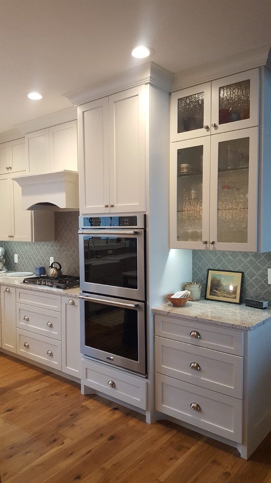 This is an example of a large traditional u-shaped eat-in kitchen in Minneapolis with a farmhouse sink, flat-panel cabinets, white cabinets, granite benchtops, grey splashback, ceramic splashback, stainless steel appliances, light hardwood floors, with island, brown floor and white benchtop.