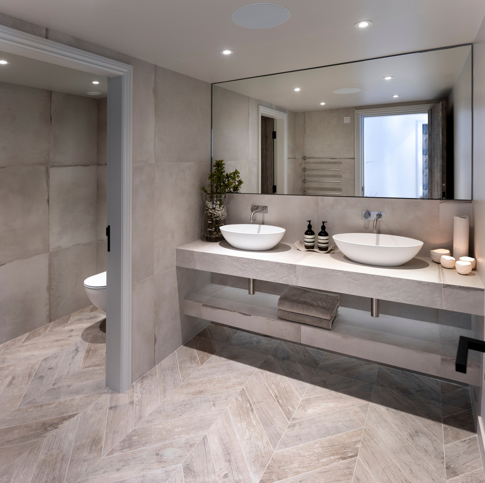 Photo of a mid-sized contemporary powder room in Cheshire with a wall-mount toilet, gray tile, porcelain tile, grey walls, porcelain floors, a trough sink, tile benchtops, grey floor and grey benchtops.