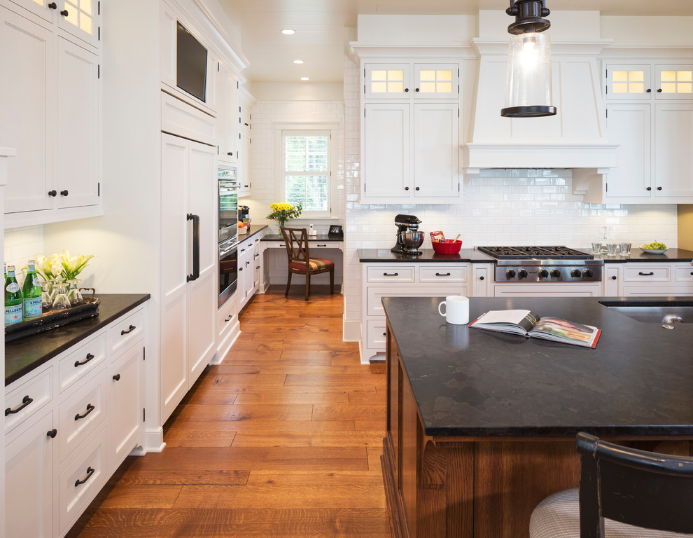 Large traditional l-shaped open plan kitchen in Minneapolis with an undermount sink, flat-panel cabinets, white cabinets, granite benchtops, white splashback, ceramic splashback, panelled appliances, with island, medium hardwood floors, brown floor and black benchtop.