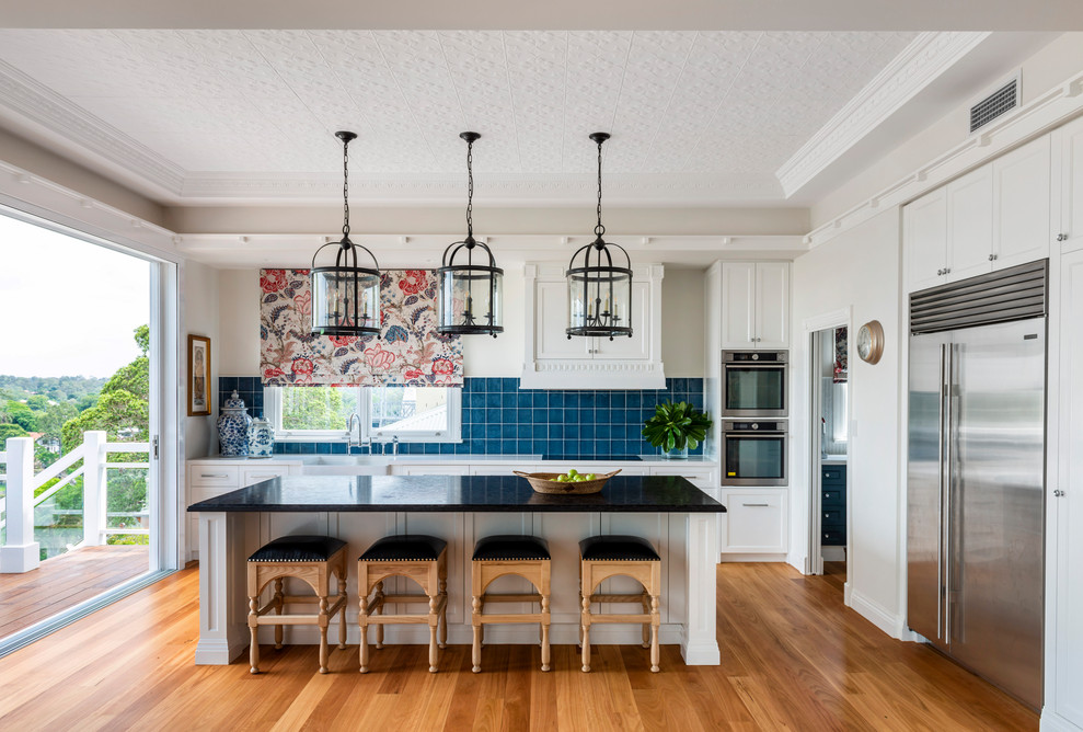 This is an example of a transitional l-shaped kitchen in Brisbane with a farmhouse sink, shaker cabinets, white cabinets, blue splashback, stainless steel appliances, medium hardwood floors, with island, brown floor and black benchtop.