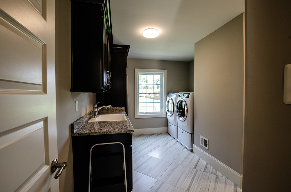 This is an example of a mid-sized traditional galley dedicated laundry room in Baltimore with a drop-in sink, recessed-panel cabinets, dark wood cabinets, granite benchtops, beige walls, porcelain floors, a side-by-side washer and dryer, grey floor and beige benchtop.