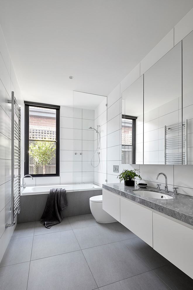 Photo of a large contemporary bathroom in Melbourne with white cabinets, a drop-in tub, a shower/bathtub combo, a wall-mount toilet, white tile, porcelain tile, white walls, porcelain floors, an undermount sink, marble benchtops, grey floor, an open shower, grey benchtops and flat-panel cabinets.