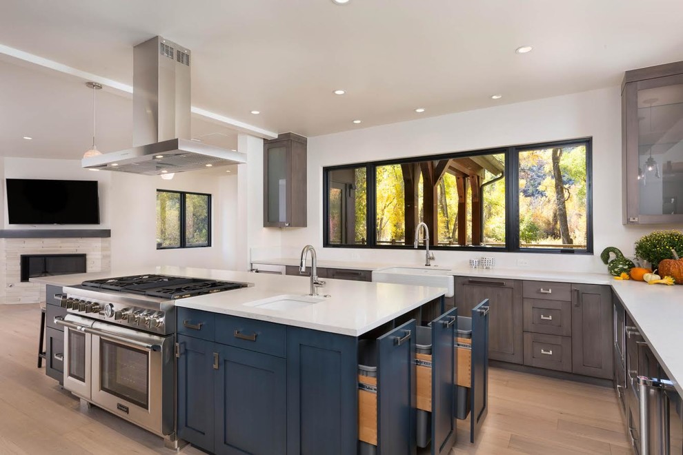 This is an example of a large transitional u-shaped open plan kitchen in Denver with blue cabinets, a farmhouse sink, recessed-panel cabinets, solid surface benchtops, stainless steel appliances, light hardwood floors, with island, brown floor and white benchtop.