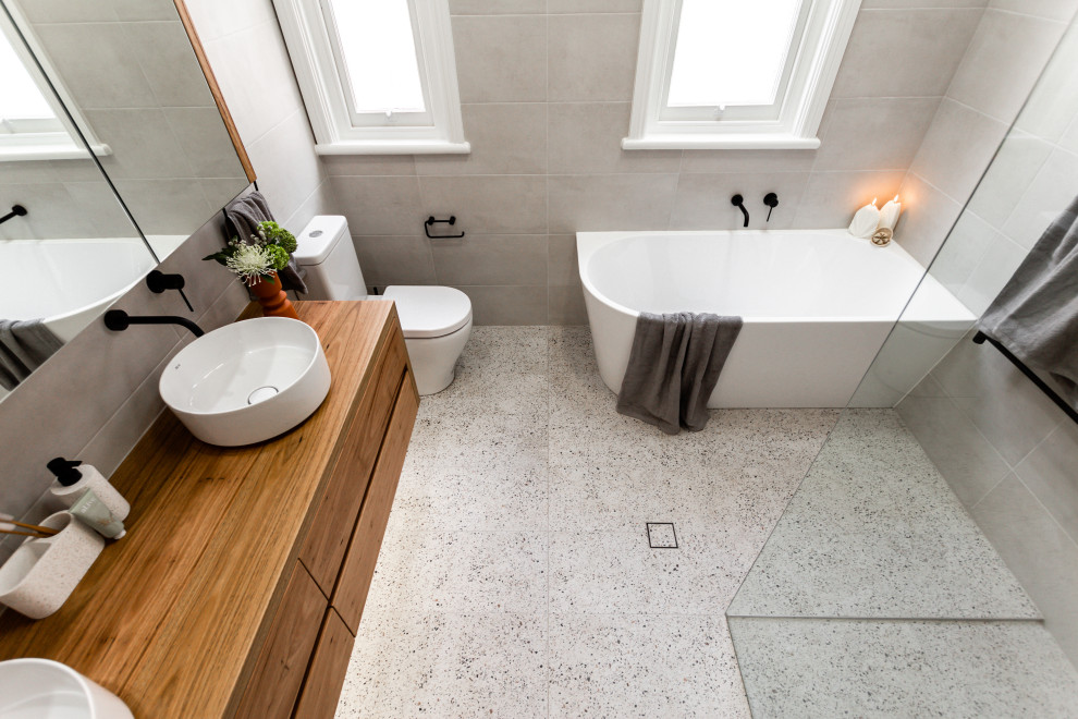 This is an example of a large modern ensuite bathroom in Sydney with flat-panel cabinets, light wood cabinets, a corner bath, a walk-in shower, a two-piece toilet, grey tiles, grey walls, terrazzo flooring, a vessel sink, wooden worktops, an open shower, double sinks and a floating vanity unit.