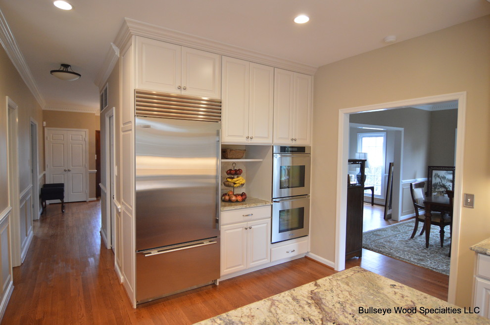 Large elegant l-shaped medium tone wood floor and brown floor kitchen photo in DC Metro with an undermount sink, raised-panel cabinets, white cabinets, granite countertops, beige backsplash, travertine backsplash, stainless steel appliances and an island