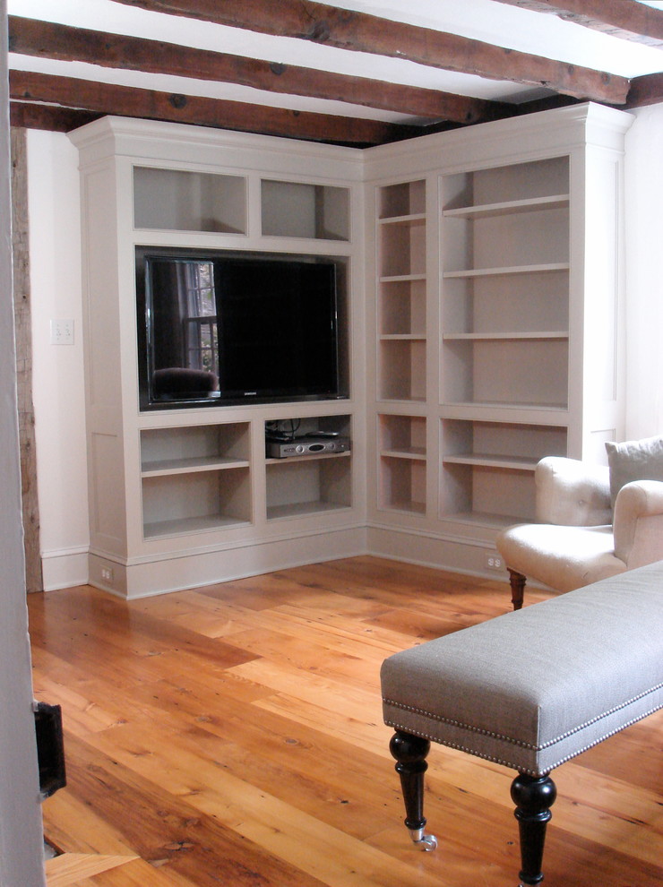 Photo of a small country enclosed living room in New York with a library, white walls, light hardwood floors, a standard fireplace, a wood fireplace surround and a wall-mounted tv.