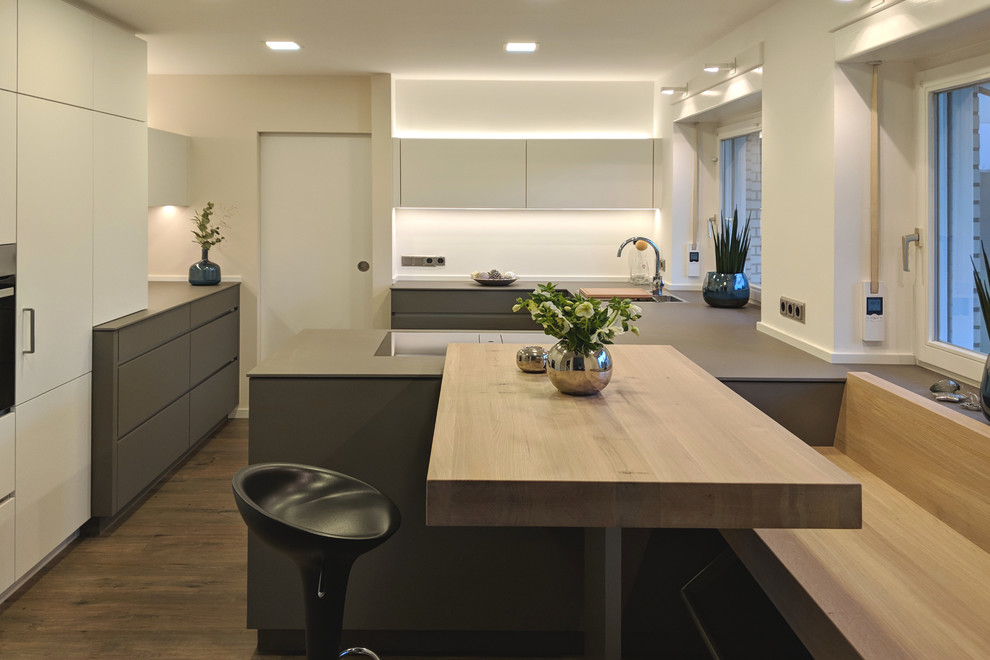 This is an example of a mid-sized contemporary u-shaped eat-in kitchen in Essen with flat-panel cabinets, white cabinets, a drop-in sink, black appliances, medium hardwood floors, multiple islands and brown floor.